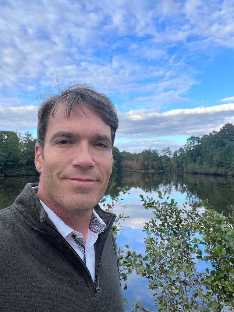 Jason Wolin outdoors by a lake, trees and sky reflected on the water