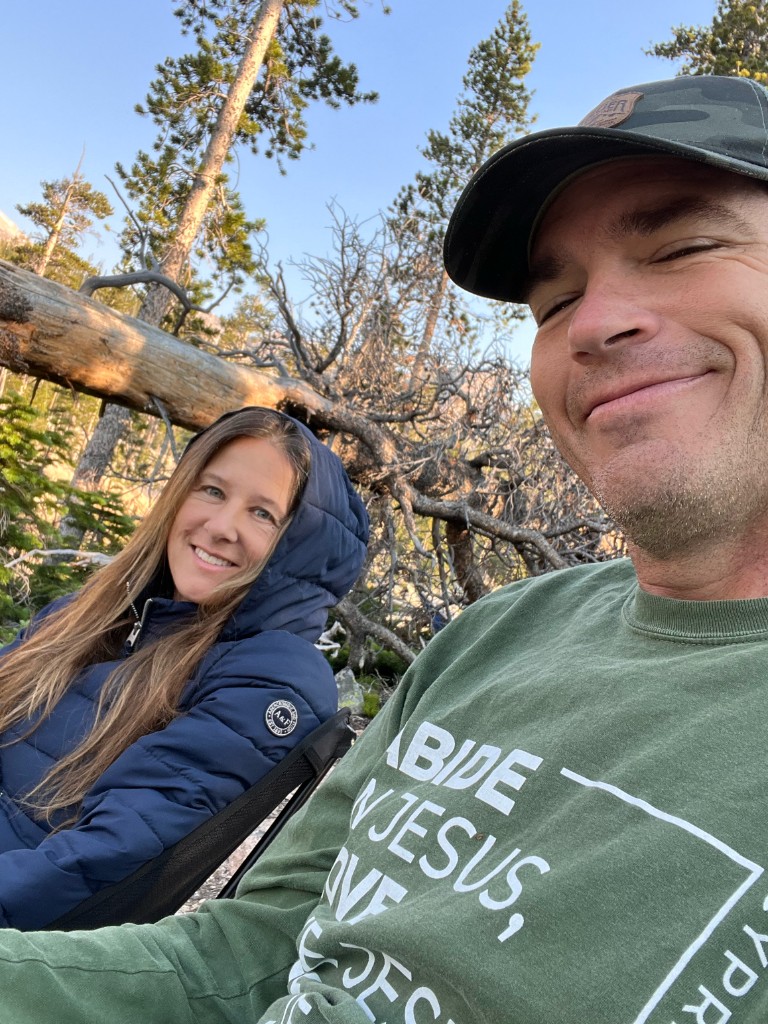 Jason and his wife outdoors in the woods, camping chairs and pine trees behind them